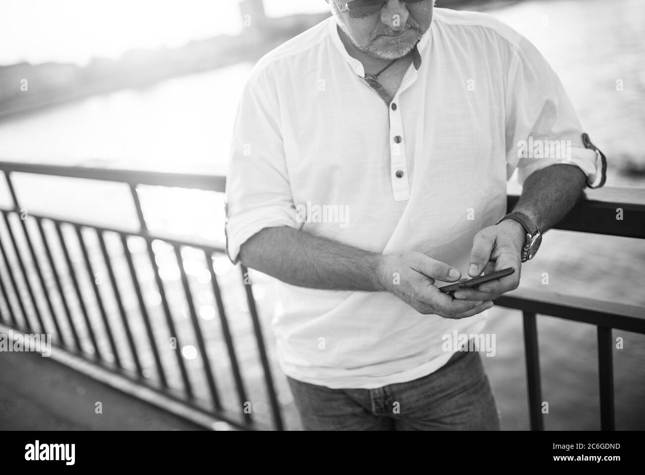 Un uomo d'affari caucasico di mezza età con occhiali, in una camicia bianca, sta digitando un messaggio sul telefono che pende contro la ringhiera del ponte. Foto Stock