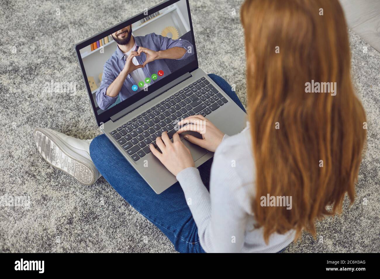 Datazione online. Segno del cuore con le mani maschili in un monitor del laptop di una ragazza giovane seduta sul pavimento a casa. Foto Stock