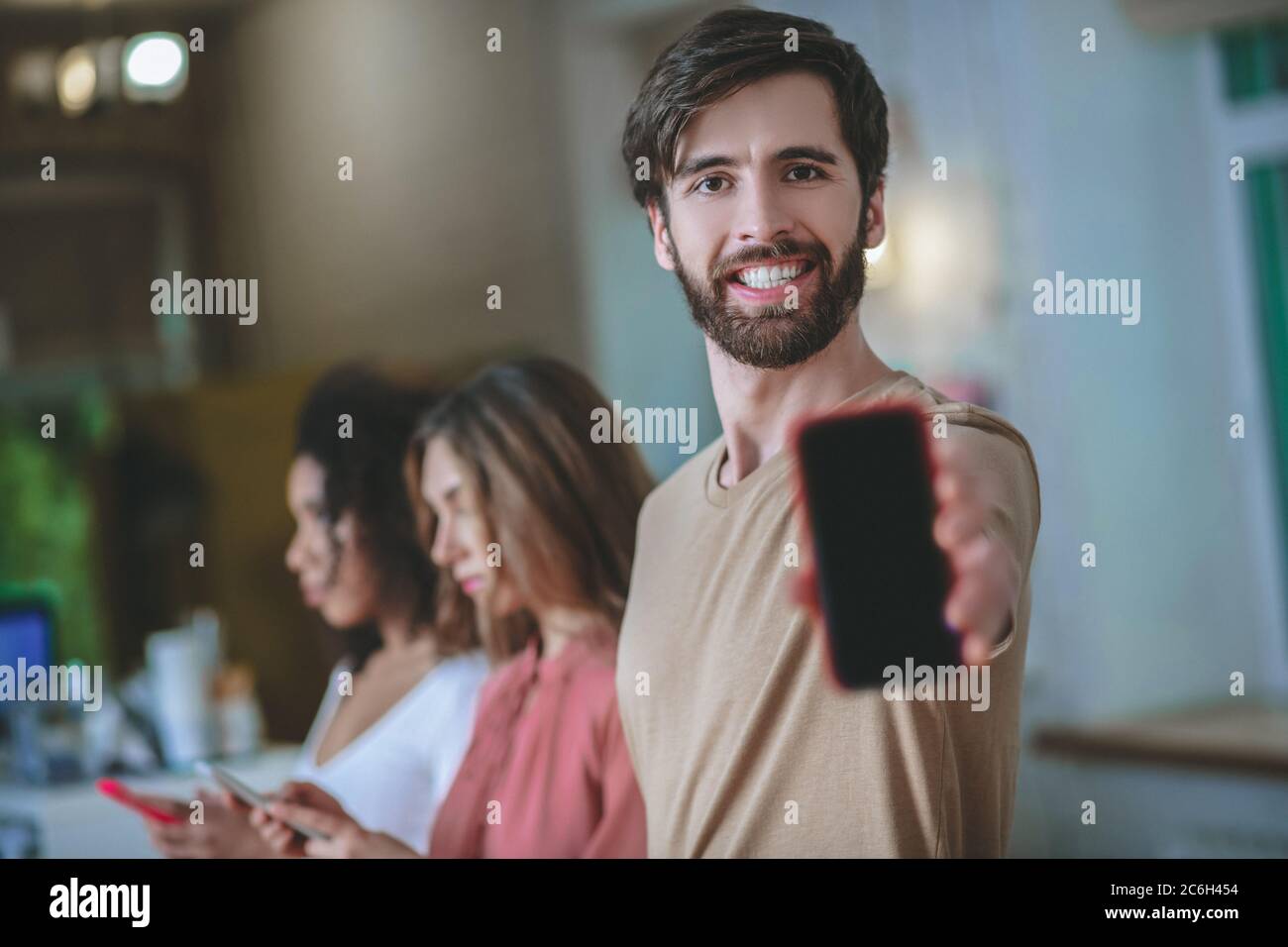 Uomo sorridente che tiene lo smartphone nel braccio allungato, due ragazze dietro Foto Stock