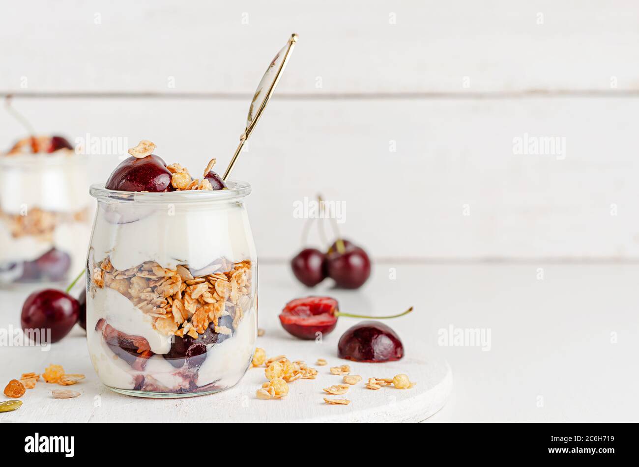 Vaso di granola con yogurt greco e ciliegie dolci su sfondo bianco di legno. Mangiare sano, spazio copia. Foto Stock
