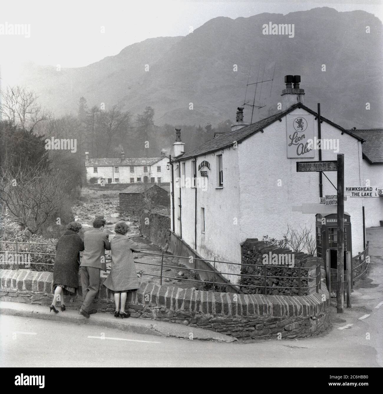 Anni '1950, storico, Coniston, Lake District, tre visitatori che guardano su un ruscello, Church Beck, accanto al Black Bull Inn, un vecchio hotel e locanda per carrozze, con un cartello Lions Ales, probabilmente non le tradizionali camminatrici distict del lago, dato che le due signore indossano scarpe con tacco. Un cartello stradale per Hawkshead e il lago. Sullo sfondo, una montagna conosciuta come "Old Man". Foto Stock