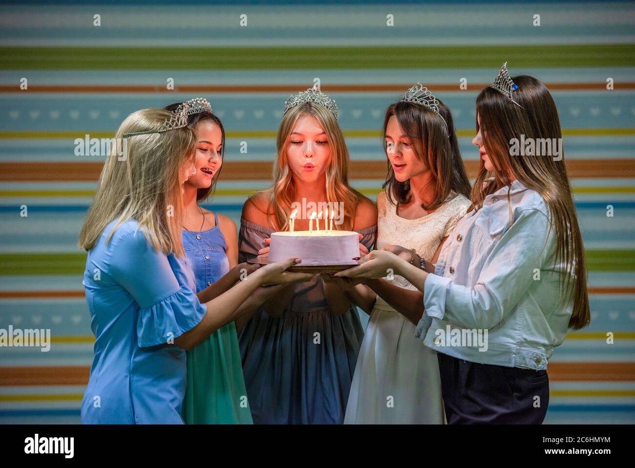 Ragazze adolescenti sorridenti graziose in abiti e corone tenendo insieme la torta e soffiando le candele alla festa di compleanno sullo sfondo colorato a strisce Foto Stock