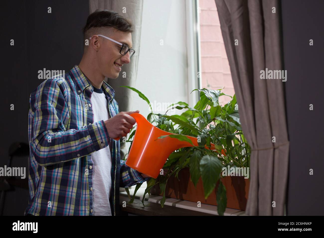 Giovane uomo che usa l'annaffiatura arancione può prendersi cura delle piante in tempo di giorno. Fiori di irrigazione freelance maschile in ufficio a casa Foto Stock