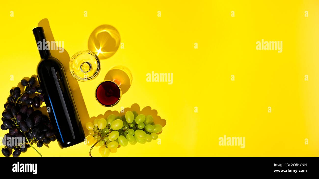 Due bicchieri di vino rosso e bianco, una bottiglia, uva su fondo giallo. Il concetto di vino italiano. Foto con ombre dure, vista dall'alto. Banner, formato lungo. Spazio libero per il testo Foto Stock