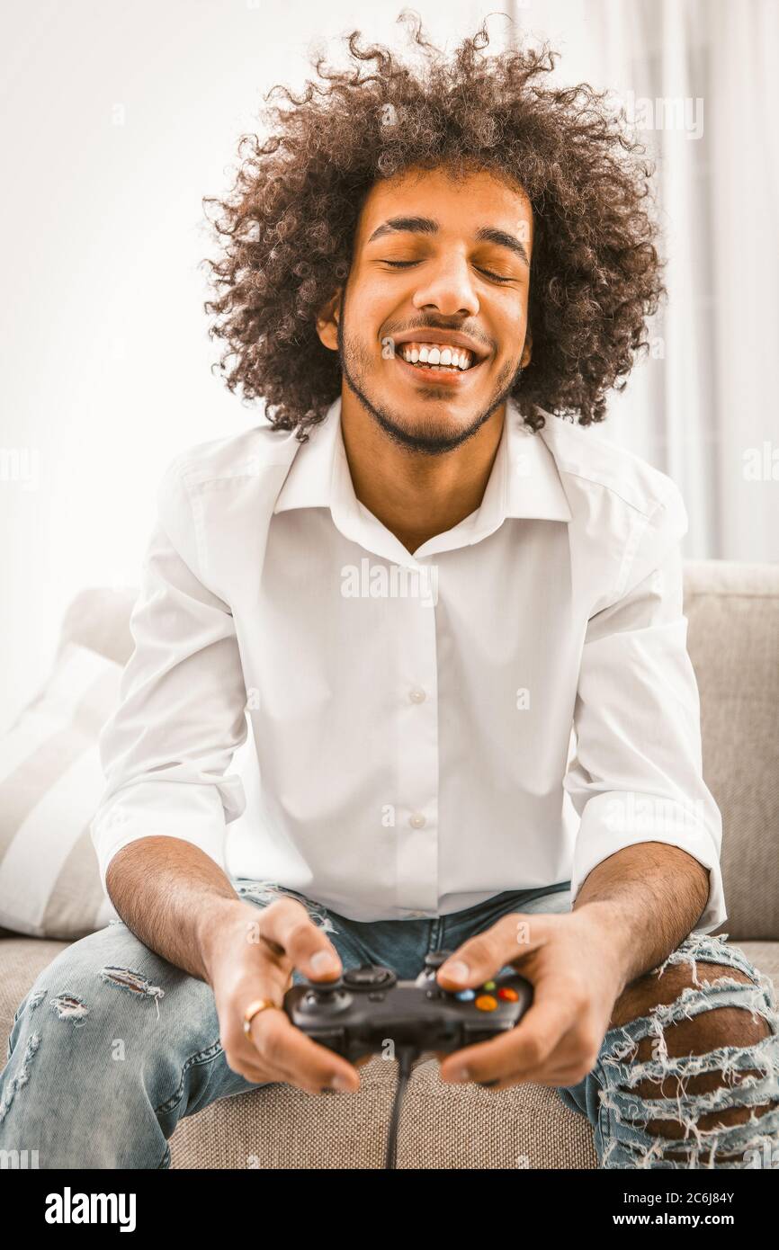 Shaggy gamer ha chiuso gli occhi con piacere e ride allegro mentre si gioca al computer, premendo i pulsanti della console di gioco. Gioco di giovane uomo Foto Stock