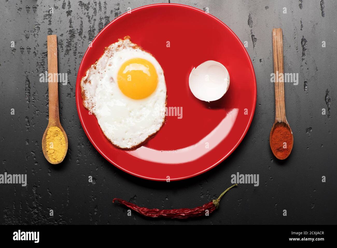 Cibo posto su piastra rossa su sfondo nero, vista dall'alto. Condire con uova fritte vicino a cucchiai con spezie e peperoncino rosso. Colazione e concetto di arte alimentare. Omelette, gusci e riflessi fanno sorridere il volto Foto Stock