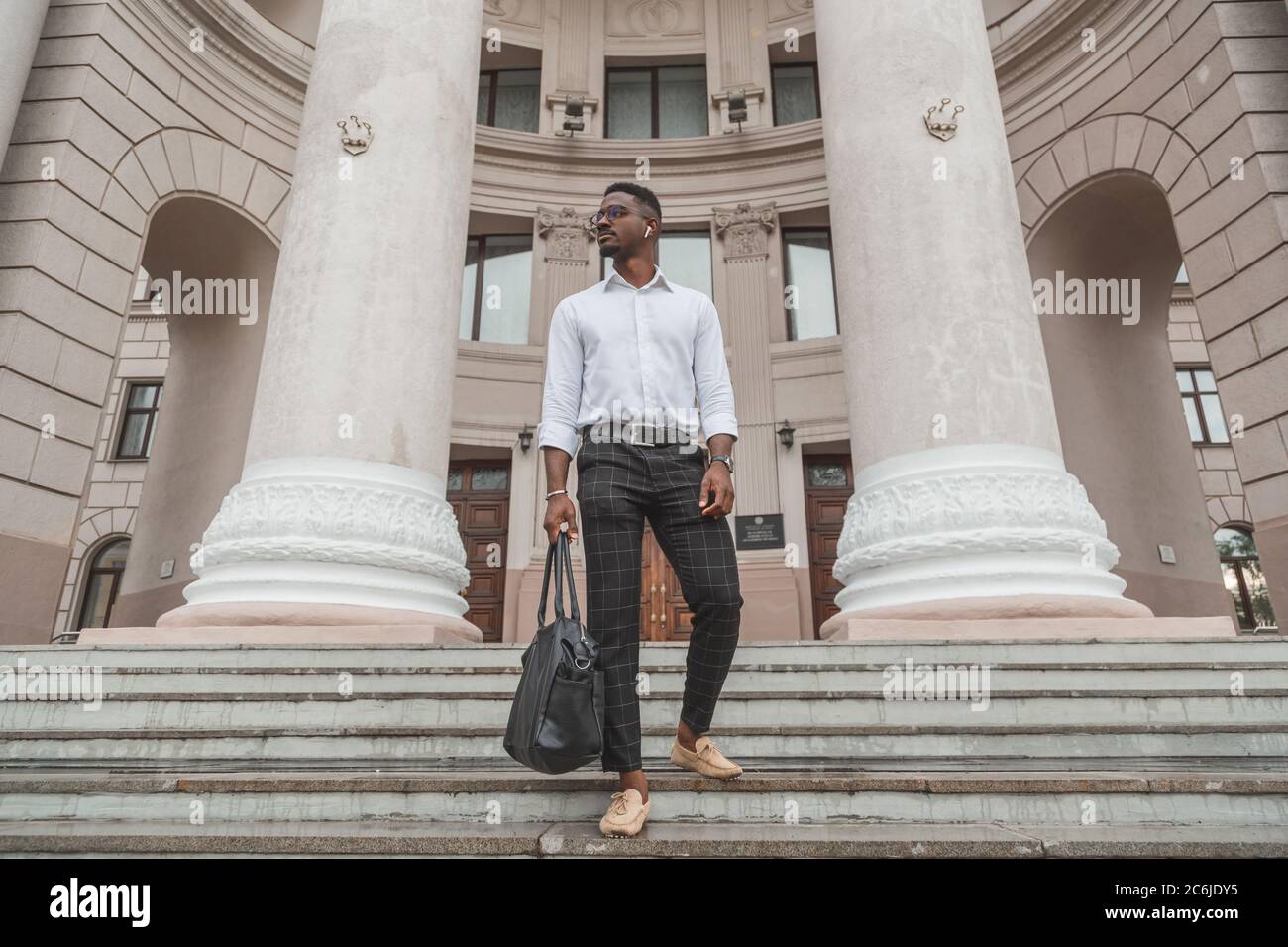 Elegante giovane uomo nero con un design elegante e auricolari wireless e borsa in pelle in mano. Foto Stock