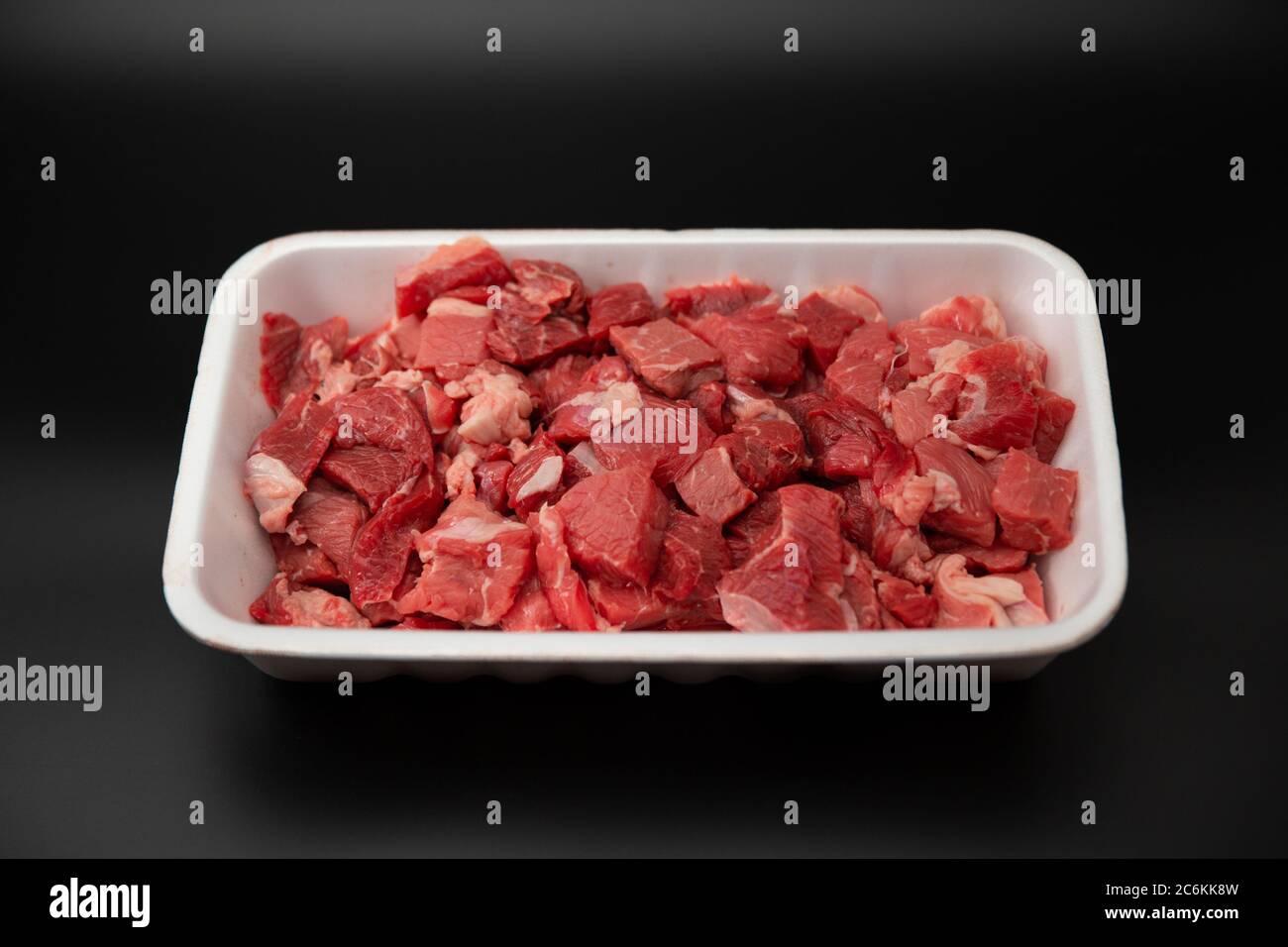 Carne grezza tagliata a cubetti in un contenitore bianco di polistirolo isolato su fondo nero con spazio per la copia del testo Foto Stock