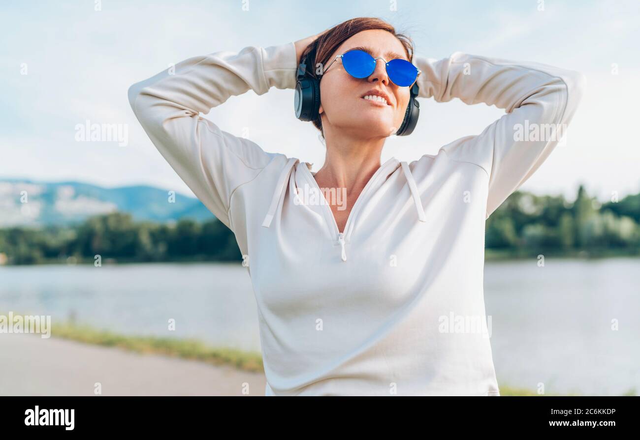 Giovane bella donna in occhiali blu da sole che ascolta la musica utilizzando smartphone e cuffie wireless sorridente allegro. Camminare con la musica preferita Foto Stock