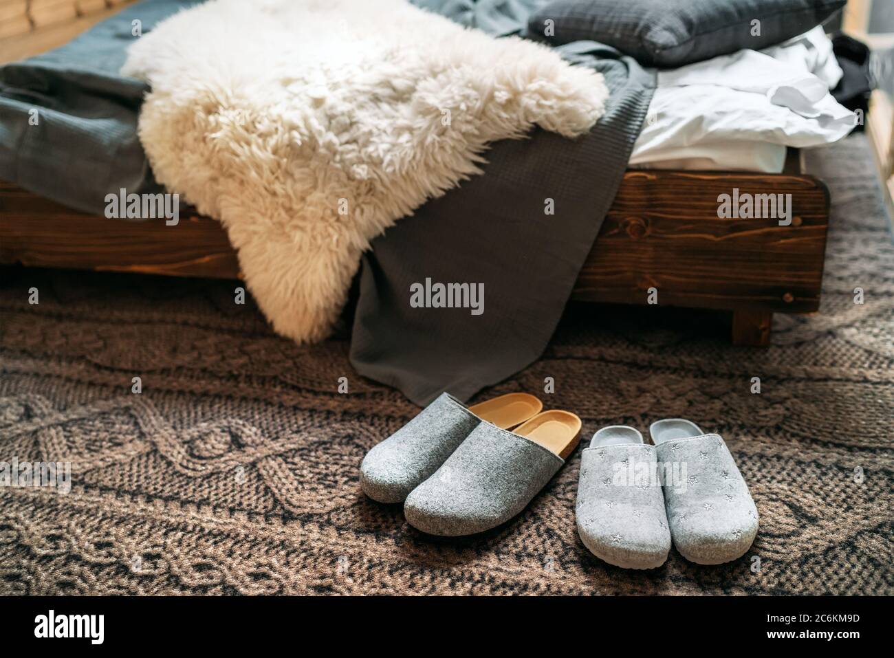 Le due pantofole grigie per la casa vicino al letto in legno sul "pavimento lavorato a maglia" nella camera da letto accogliente. Home dolce casa concetto immagine. Foto Stock