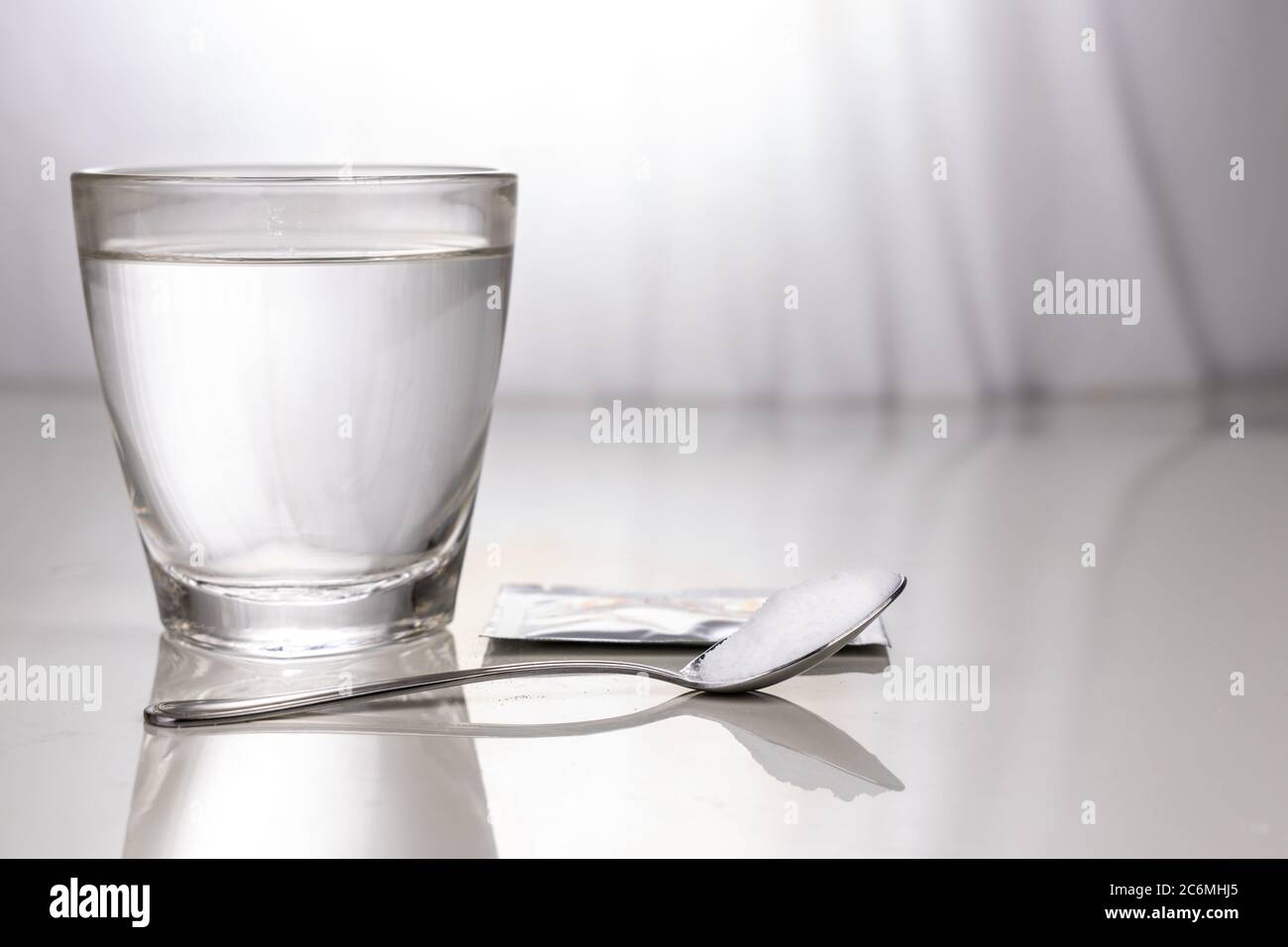 Sale di reidratazione orale o orale con un bicchiere d'acqua, bustina e cucchiaio sul tavolo contro la luce della finestra Foto Stock