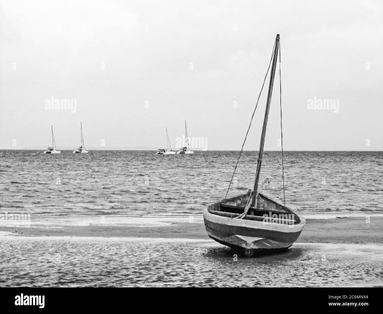 Un piccolo dhow in legno sulla spiaggia di Vilanculos, Mozambico, al mattino presto, con quattro yacht a vela sullo sfondo, fotografati in Monocromo Foto Stock