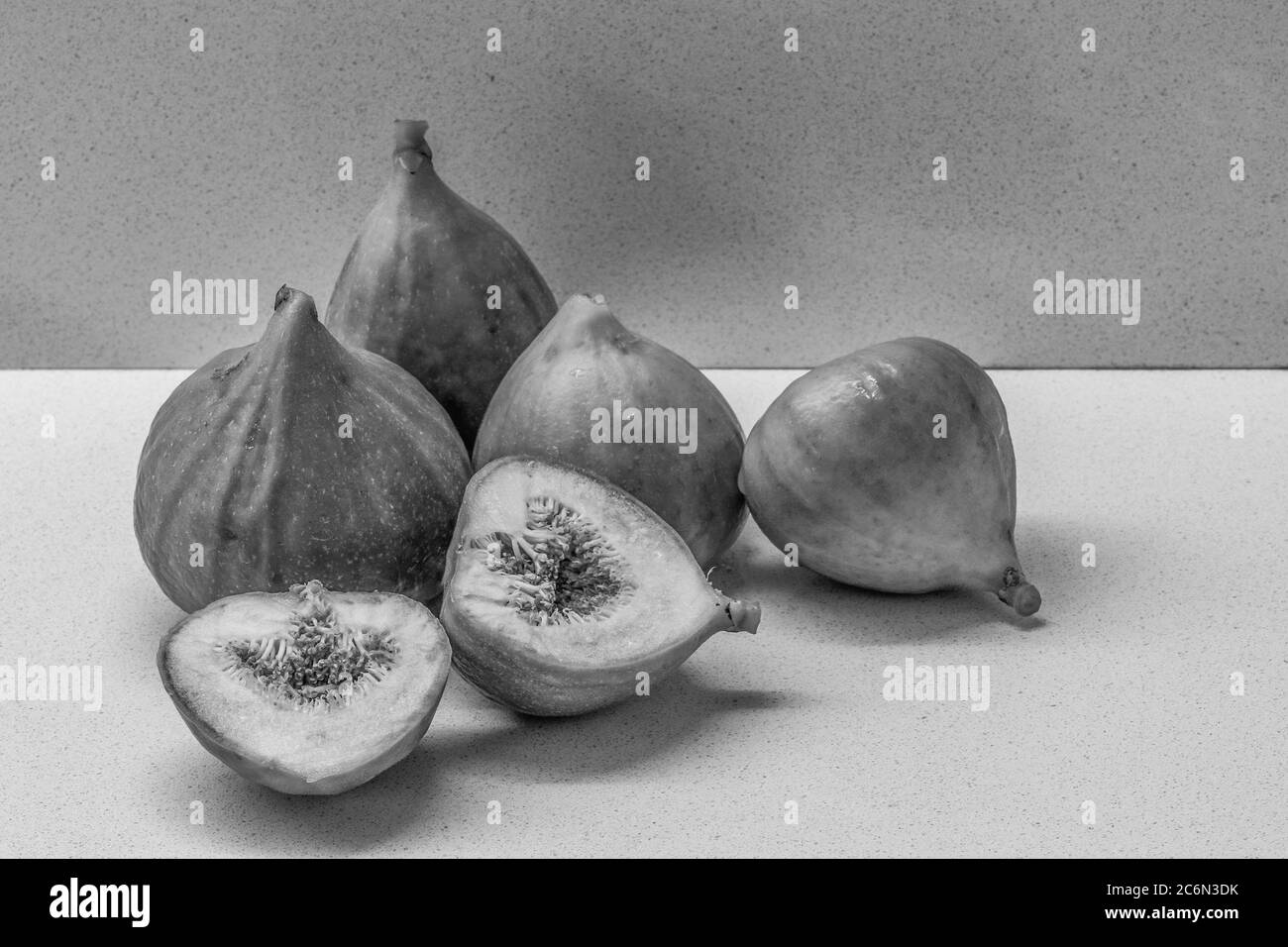 Un primo piano bianco e nero di alcuni fichi maturi, uno dei quali è tagliato a metà, su una superficie grigio chiaro. Foto Stock