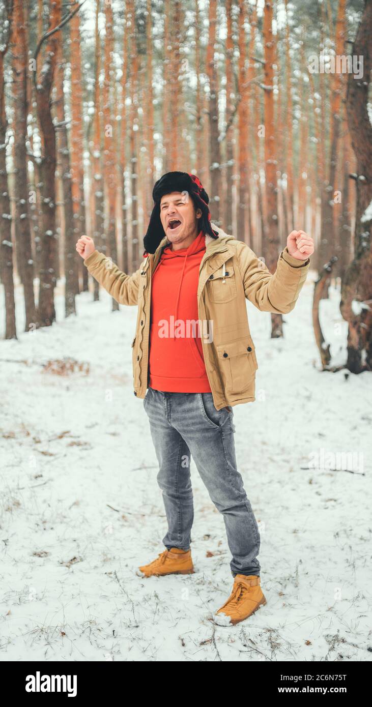 Bell'uomo in piedi e che canta in foresta nella stagione invernale. Giovane maschio che indossa una giacca beige sopra una felpa con cappuccio rossa e un cappello con le braccia Foto Stock