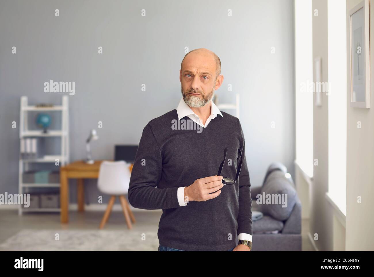 Ritratto di un uomo anziano che guarda la fotocamera a casa. Uomo serio maturo in piedi con gli occhiali in mano in interni Foto Stock