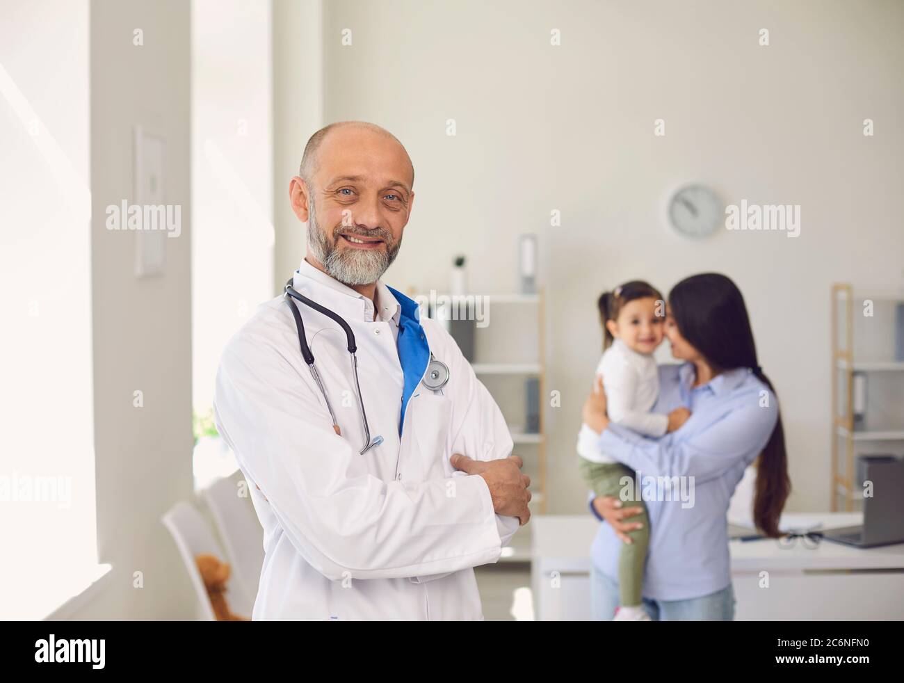 Sorridente medico pediatrico in studio medico, con bambina e madre in background. Foto Stock