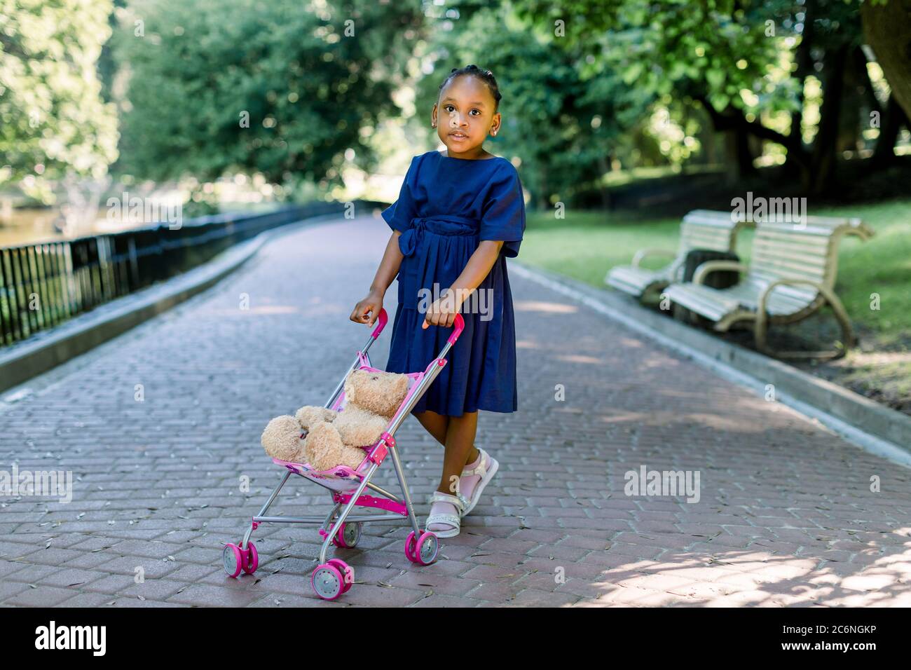 Piccola bambina africana di 5 anni cammina e gioca con il suo passeggino giocattolo nel parco. Carino bambina scura con un gramolo e un orsacchiotto Foto Stock