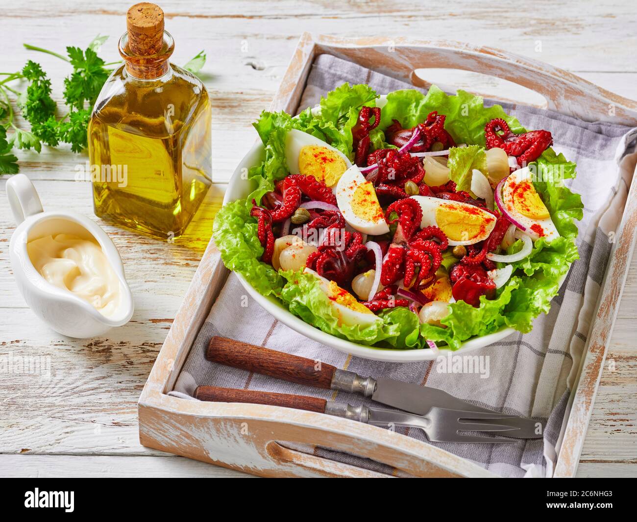 primo piano di insalata di polpo con uova sode, capperi, cipolla marinata su foglie di lattuga in una ciotola su un vassoio, su un rustico tavolo di legno con oli Foto Stock