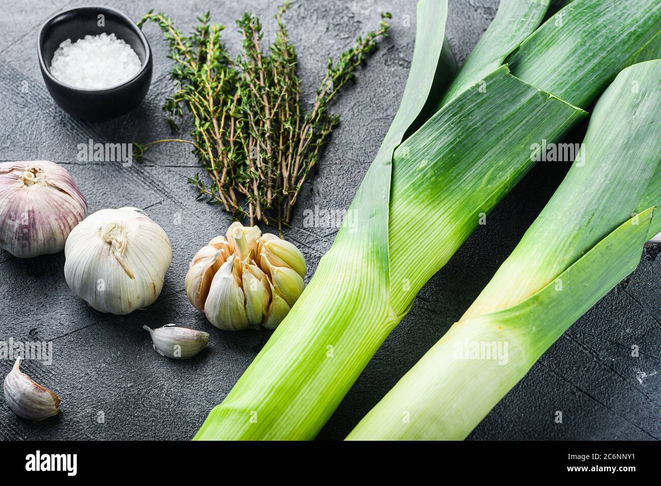 Gambi di porro con erbe ingredienti per cucinare porri brasati, su tavola grigia testurizzata vista laterale. Foto Stock