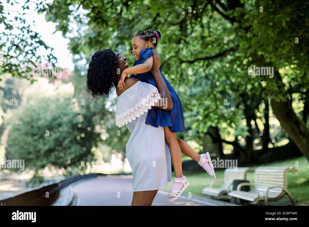 Giovane bella donna africana madre in azzurro vestito giocando con la sua piccola figlia carina nel parco, divertendosi e sollevando il bambino. Felice Foto Stock