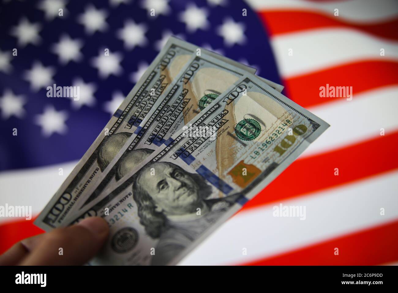 Visualizza su mano isolata tenendo tre banconote da 100 dollari. Sfondo della bandiera USA sfocato. (Attenzione alla fattura corretta "States") Foto Stock