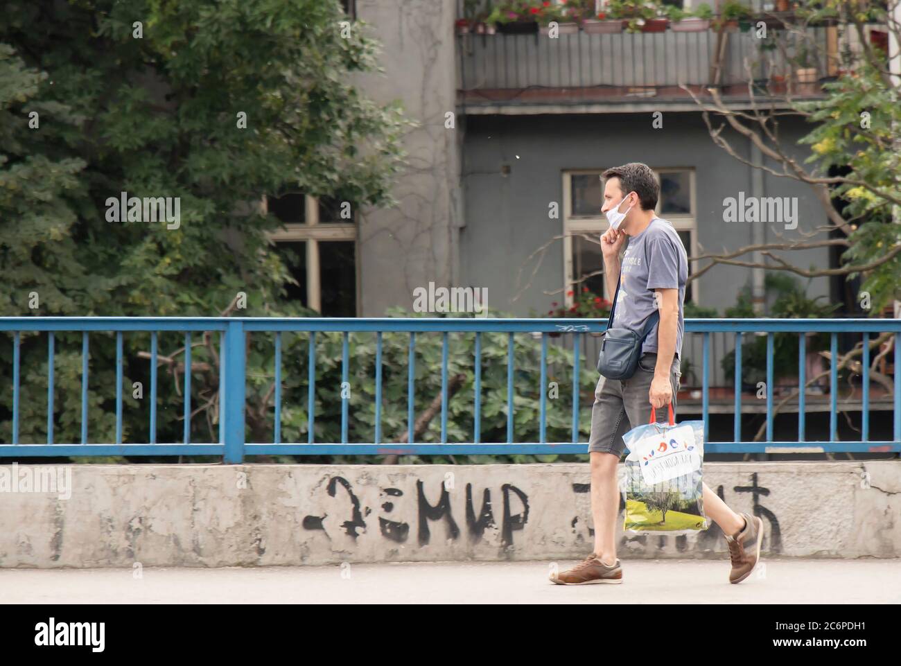 Belgrado, Serbia - 3 luglio 2020: Un uomo che indossa una maschera chirurgica a piedi lungo la strada con borsa della spesa accanto all'edificio residenziale in estate Foto Stock