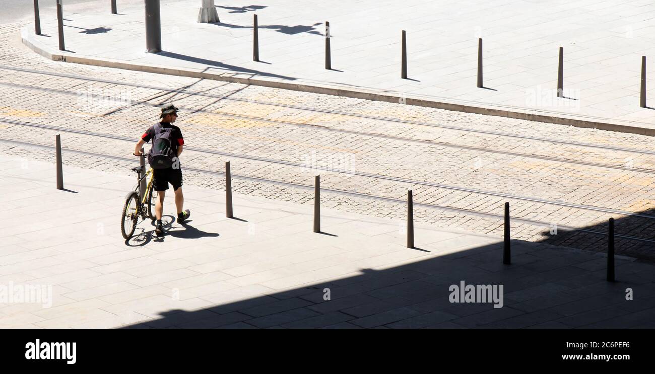 Belgrado, Serbia - 9 luglio 2020: Un giovane uomo che cammina accanto alla sua bicicletta mentre attraversa la strada con il tram in una calda giornata estiva, posteriore ad angolo alto Foto Stock