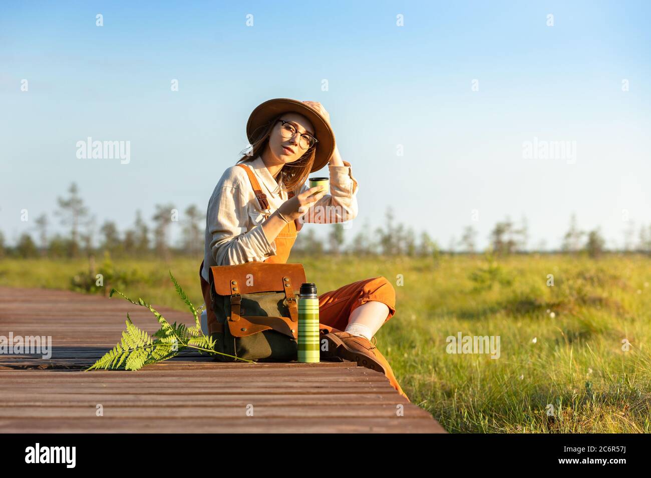 Donna botanica con zaino seduto su un percorso di legno attraverso palude torba nel parco nazionale della fauna selvatica. Naturalista riposante sul lungomare, bere tè, e. Foto Stock