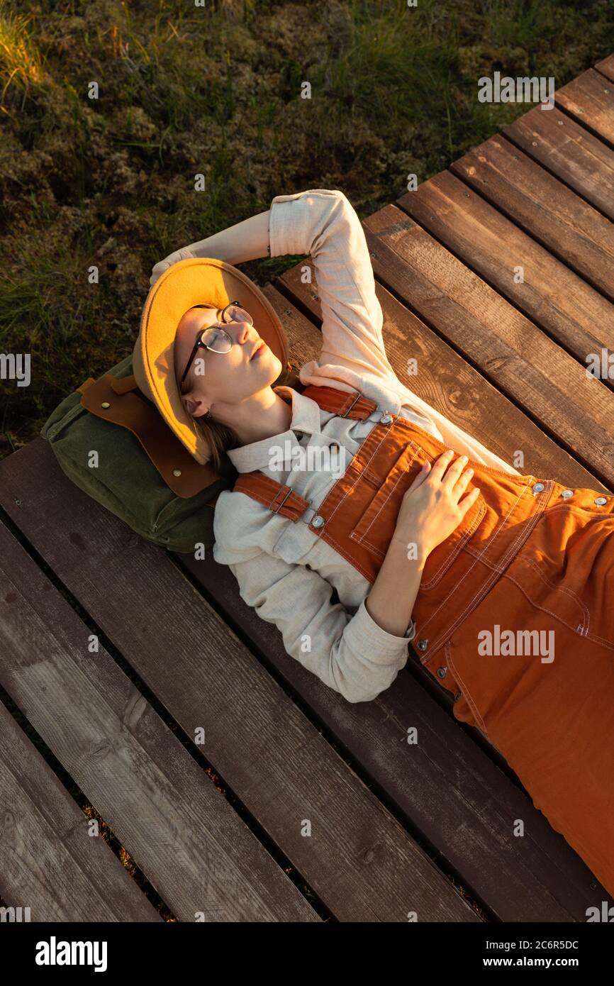 Donna naturalista in cappello beige e tute arancioni poggianti su uno zaino su un percorso di legno attraverso palude torba nel parco nazionale della fauna selvatica a sole Foto Stock