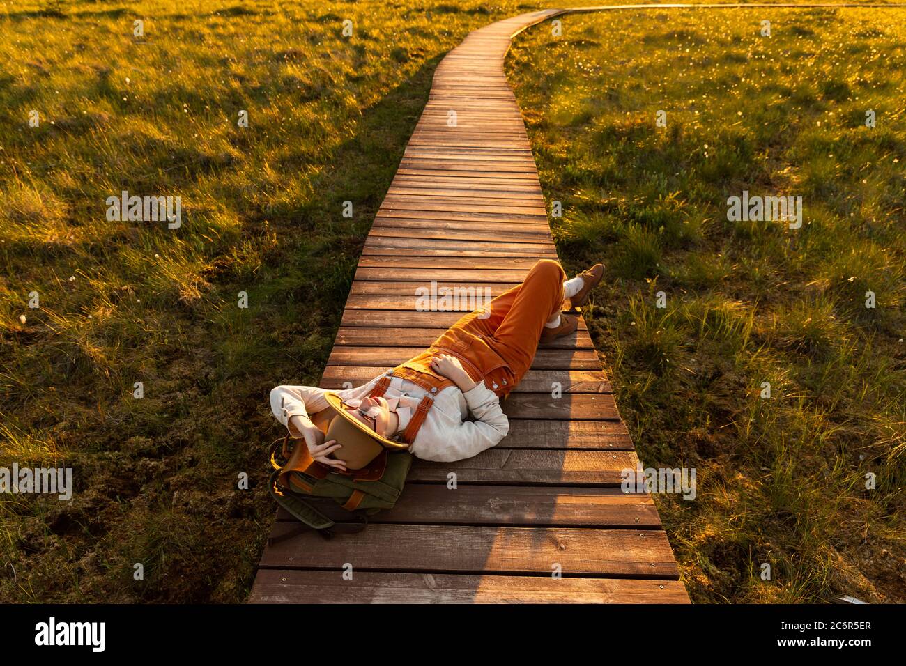 Donna naturalista in cappello beige e tute arancioni poggianti su uno zaino su un percorso di legno attraverso palude torba nel parco nazionale della fauna selvatica a sole Foto Stock