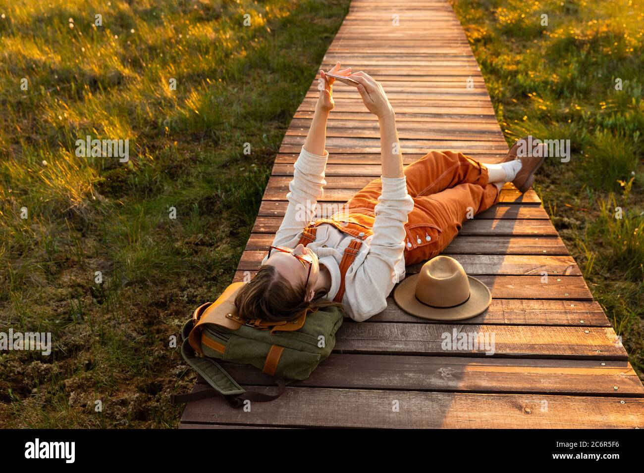 Donna naturalista arancione tute rende selfie al telefono, riposando su zaino su un percorso di legno attraverso palude torba in fauna selvatica parco nazionale Foto Stock