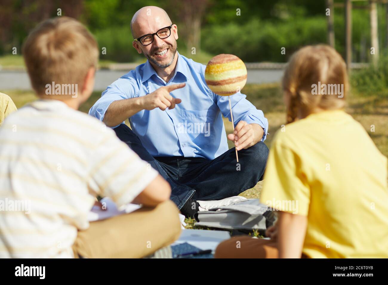 Ritratto di insegnante maschile calvo che punta al modello del pianeta e sorridente mentre si parla con il gruppo di bambini durante la lezione all'aperto in luce del sole, copia spazio Foto Stock