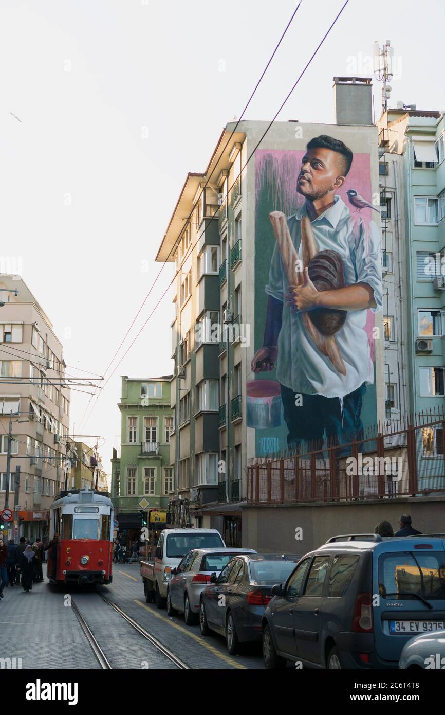Istanbul, Turchia - 23 febbraio 2020: Una grande arte murale di un giovane uomo sta tenendo il pane a Istanbul Moda. Un nostalgico tram aspetta i passeggeri a. Foto Stock