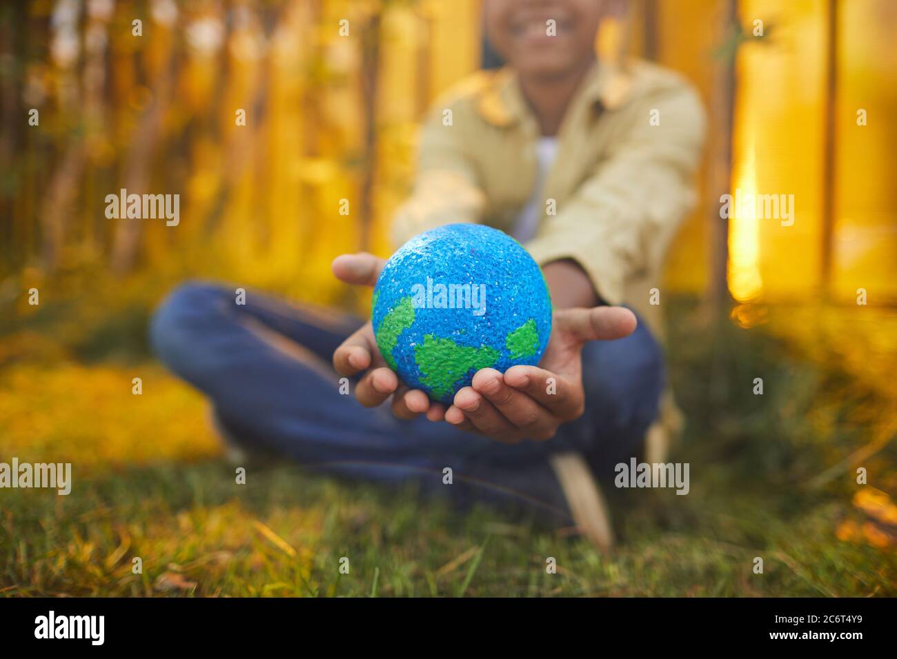 Primo piano di un ragazzo afroamericano irriconoscibile che tiene il pianeta in mano mentre si siede su erba verde all'aperto alla luce del sole, copia spazio Foto Stock