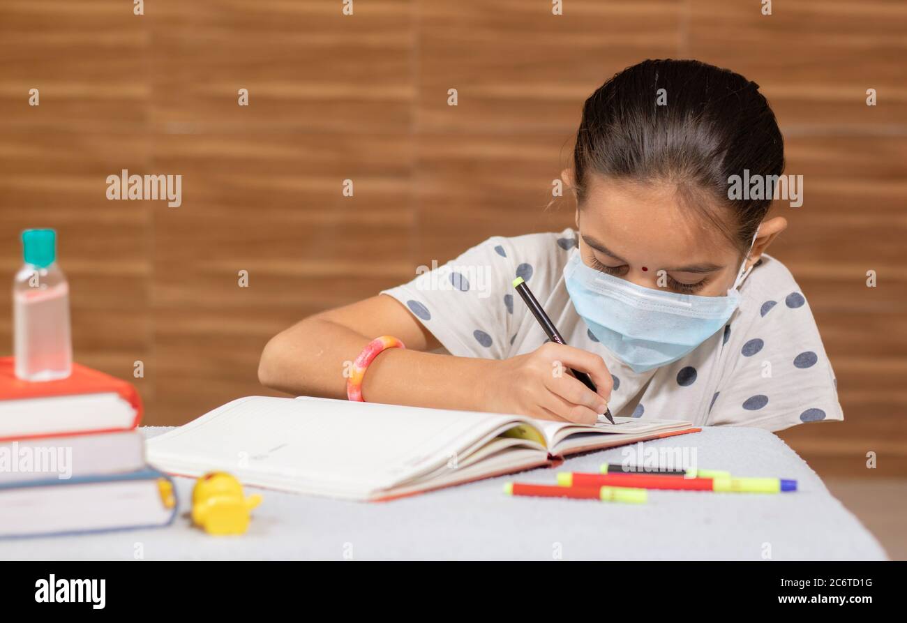 Concetto di homeschooling, ragazza giovane con maschera medica occupato nella scrittura a casa durante il covid-19 o pandemia di coronavirus. Foto Stock