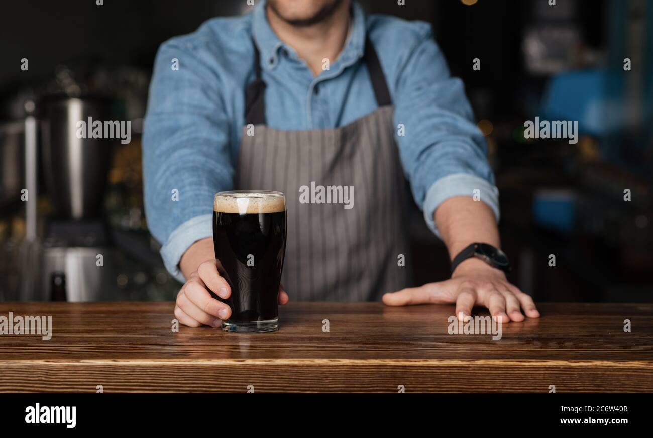 Barman in camicia e grembiule in denim si erge dietro il bancone di legno e dà un bicchiere di birra scura Foto Stock