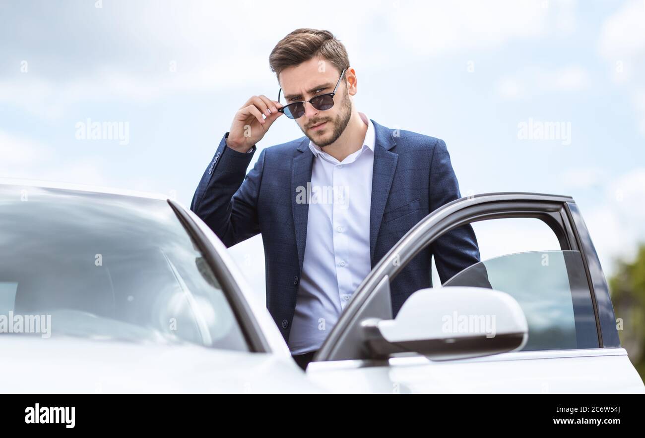 Direttore generale serio in abbigliamento formale e occhiali da sole che entrano nella sua automobile nel centro della zona della città moderna Foto Stock
