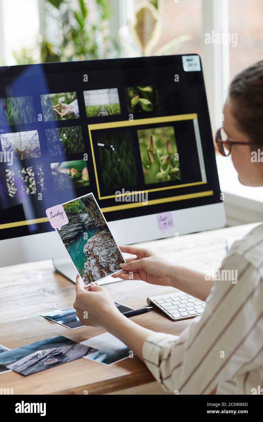 Ritratto verticale di una giovane fotografa femminile che tiene foto e sorride mentre si siede alla scrivania con un software di editing fotografico sullo schermo del computer Foto Stock