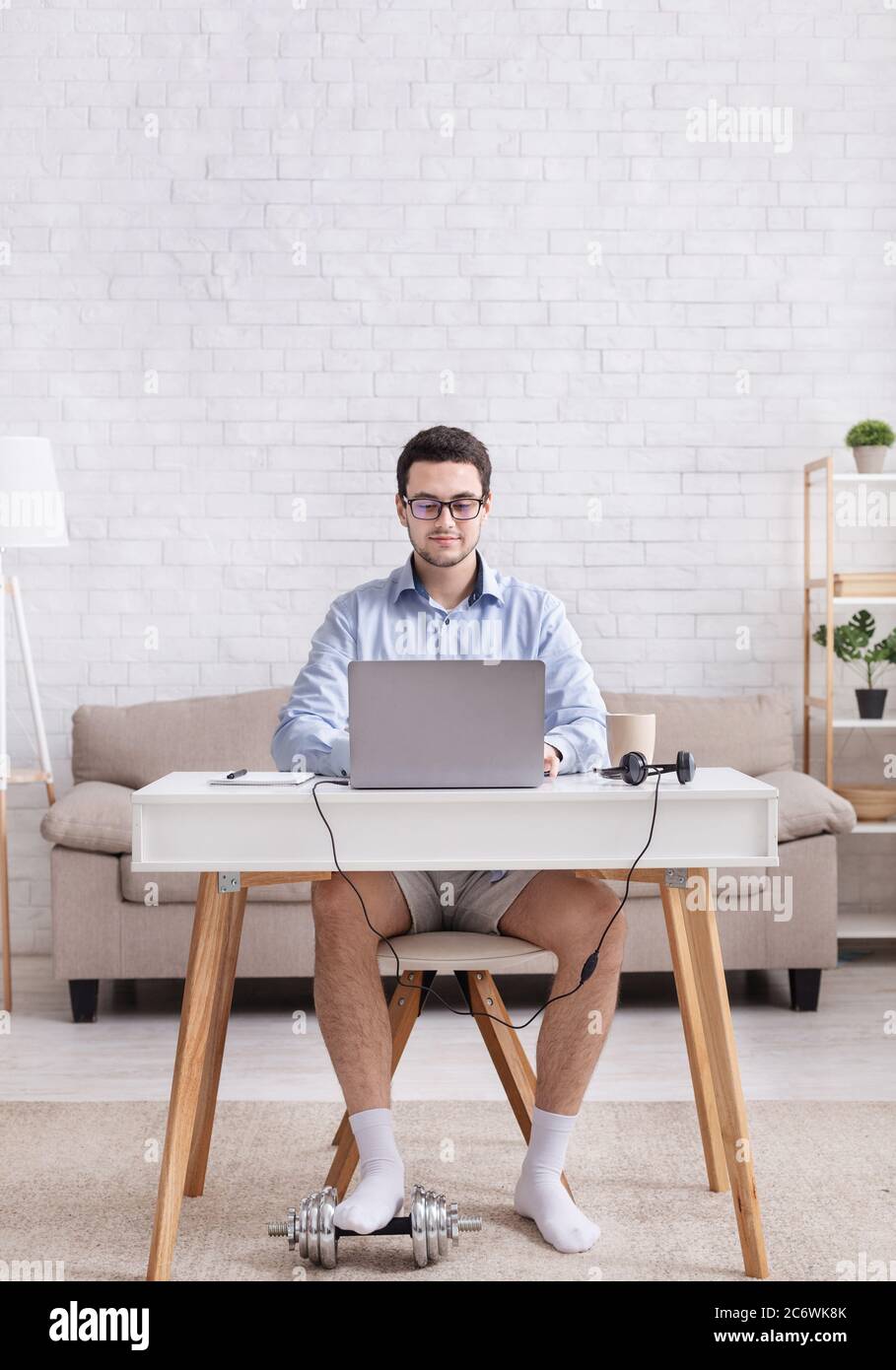 Lavoro da casa e umorismo. Un ragazzo impegnato e serio in camicia e shorts lavora al computer portatile e mette piede sul manubri Foto Stock