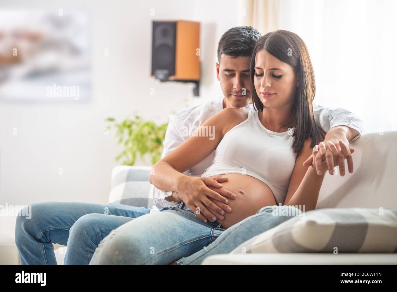 Una coppia felice che aspetta un bambino si siede su un divano, con entrambe le mani che tengono sul ventre. Foto Stock