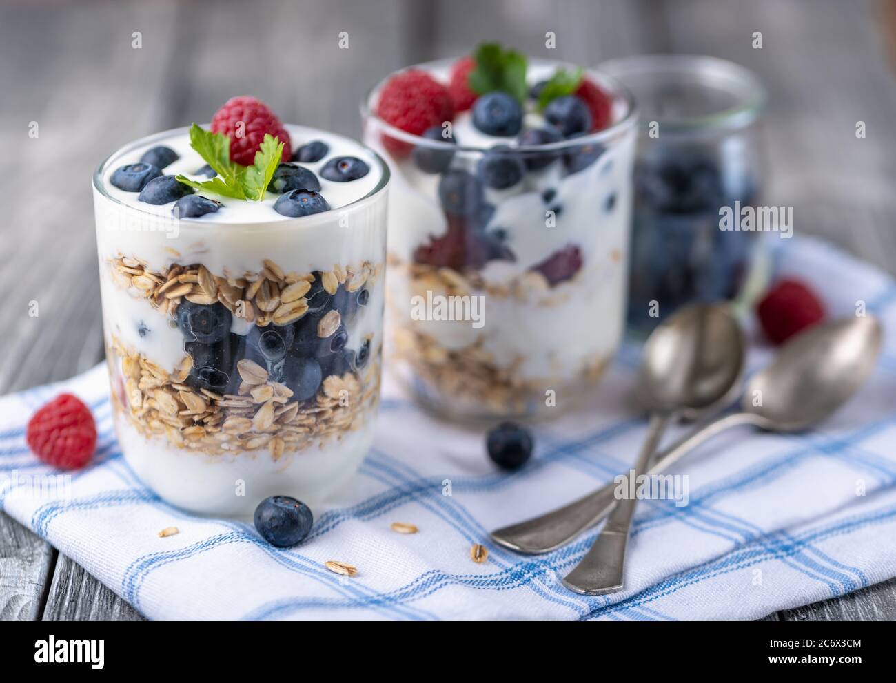 Granola con yogurt bianco con lamponi e mirtilli in due bicchieri su tovagliolo blu. Foto Stock