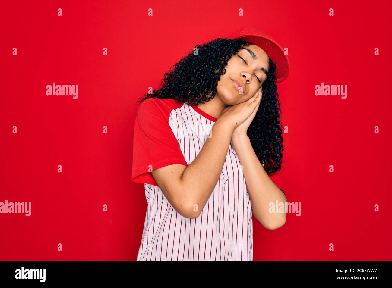 Giovane sportivo africano-americano con cappello da baseball e t-shirt a righe che dormono stanchi sognando e posando con le mani insieme mentre sorride Foto Stock