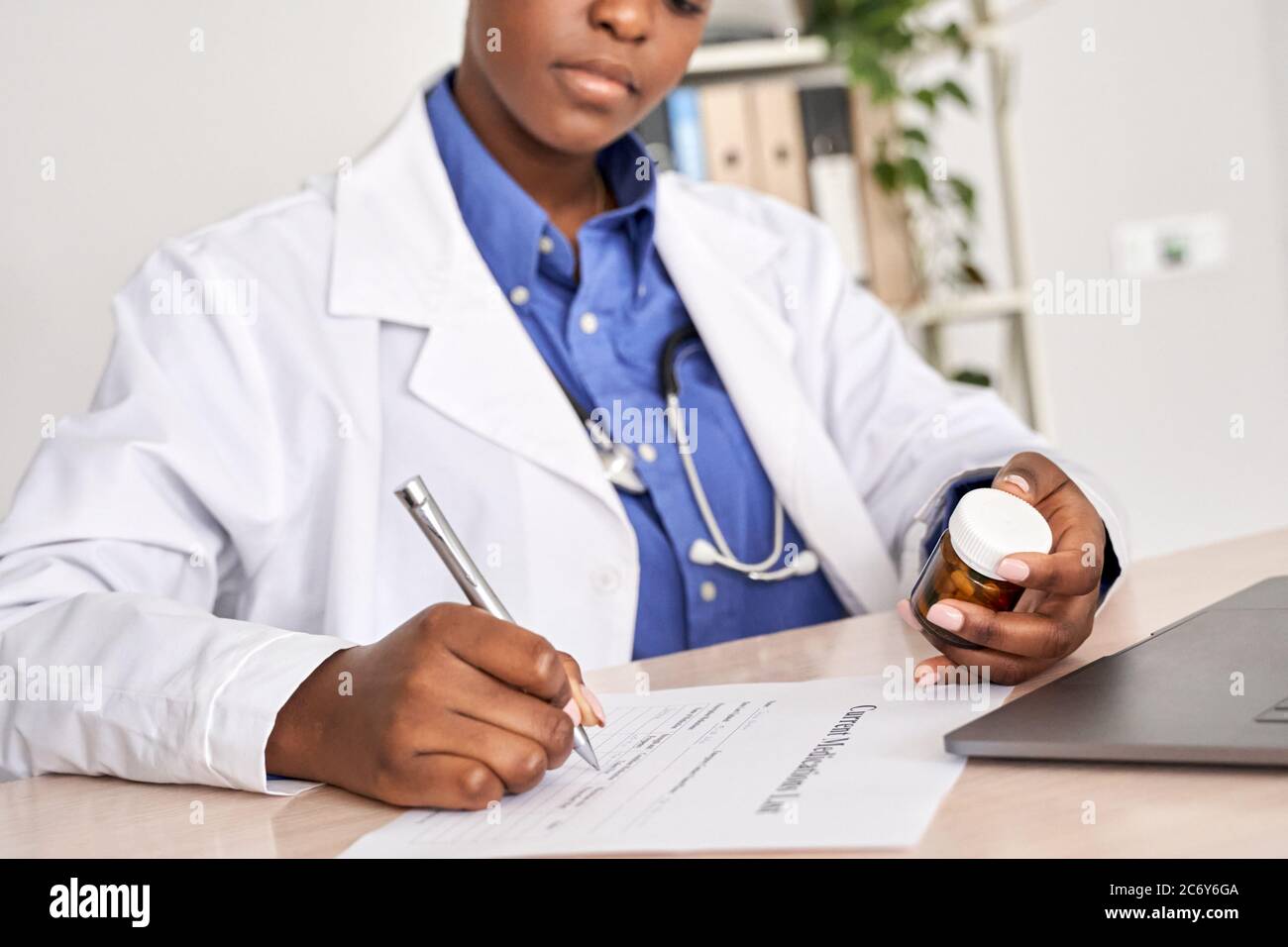 Medico afroamericano che scrive la prescrizione che tiene le pillole della bottiglia siedono alla scrivania. Foto Stock