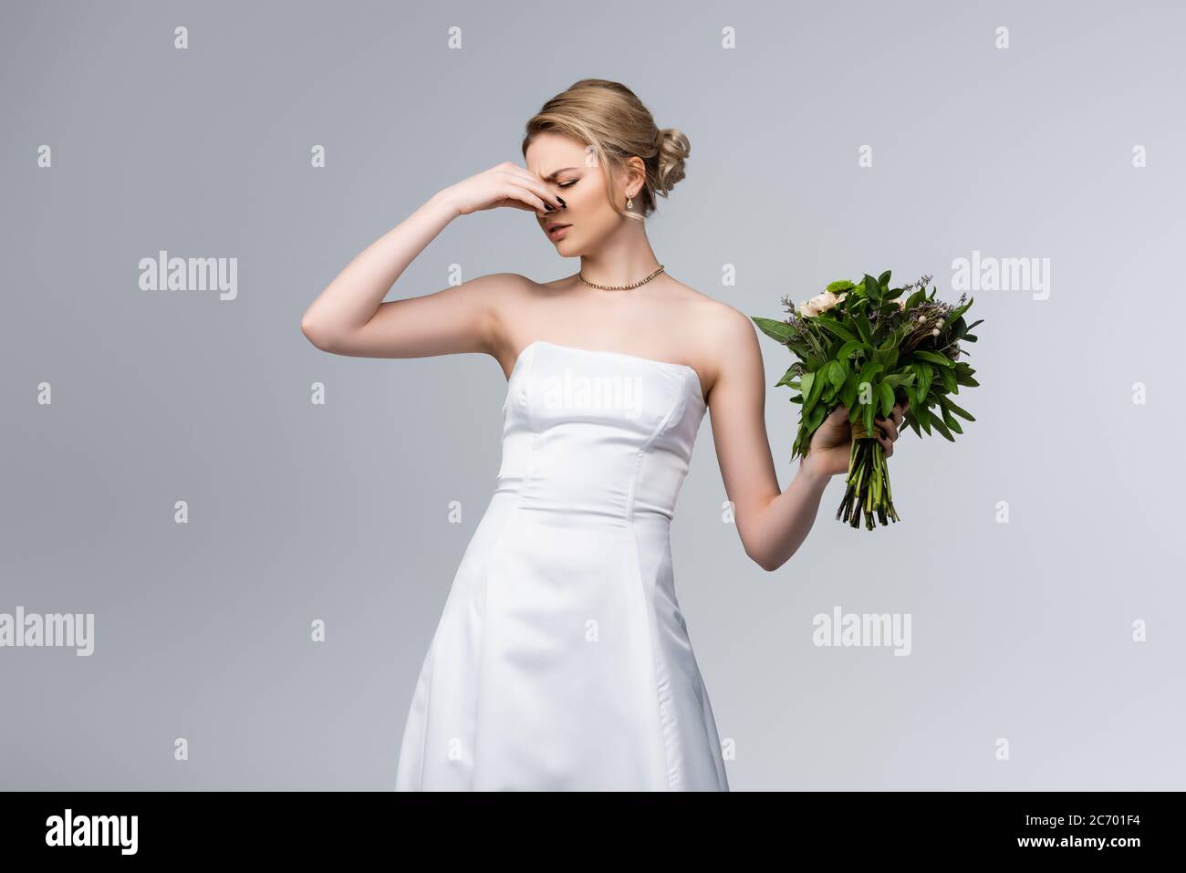 sposa dispiaciuta in abito da sposa bianco che tocca il naso mentre tiene i fiori puzzolenti isolati su grigio Foto Stock