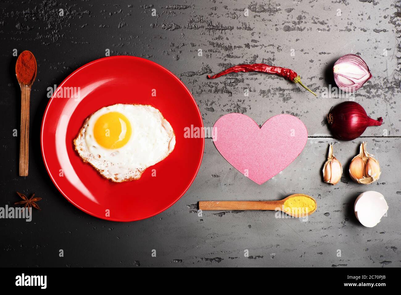 Cibo posto su piatto rosso su sfondo scuro. Omelette messa al centro del piatto. Colazione e concetto di arte alimentare. Condire con l'uovo vicino a cucchiai con spezie, aglio, cipolla, gusci di uovo, peperoncino e cuore rosa Foto Stock