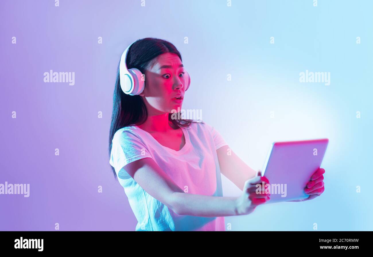 I dispositivi moderni sono sorpresi. Scioccata ragazza asiatica in cuffie bianche guarda tablet con schermo luminoso Foto Stock