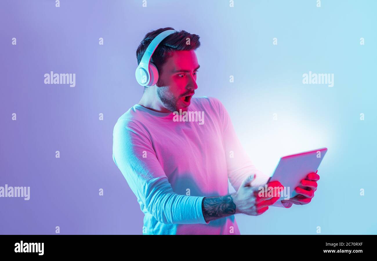 Sorprendente online. L'uomo scioccato con la bocca aperta in cuffia trattiene la luce al neon del tablet Foto Stock