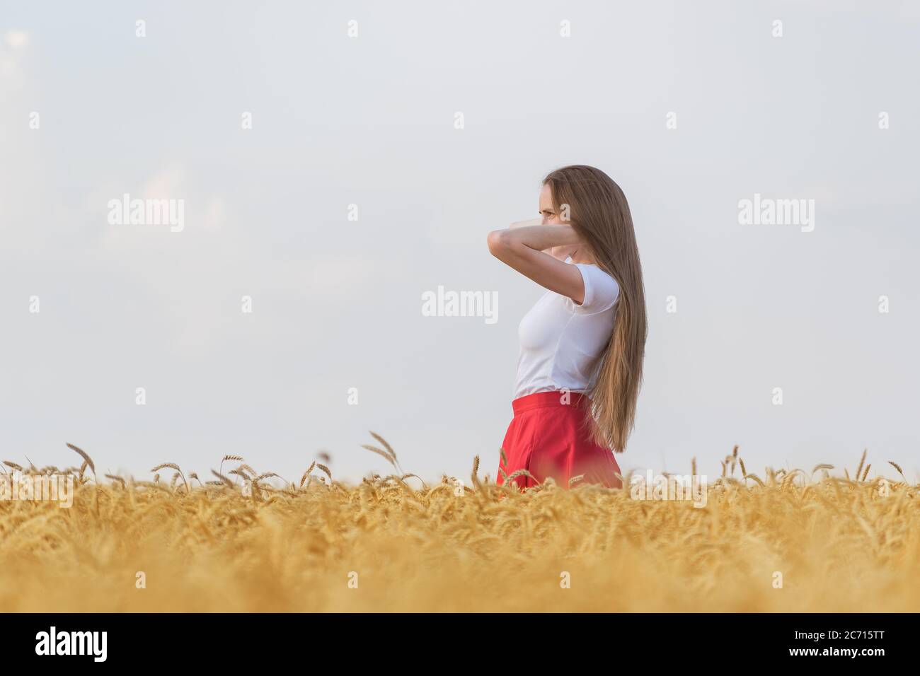 Bella donna giovane con lunghi capelli belli in piedi tra stocchi di grano maturo. Vista laterale. Foto Stock