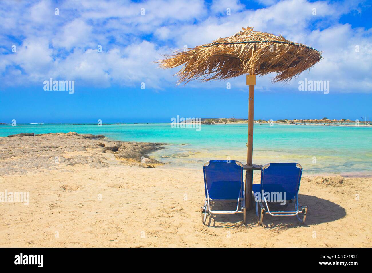 Due lettini blu e ombrellone su spiaggia di sabbia rosa con rocce, acqua turchese e nuvole nel cielo blu. Foto Stock