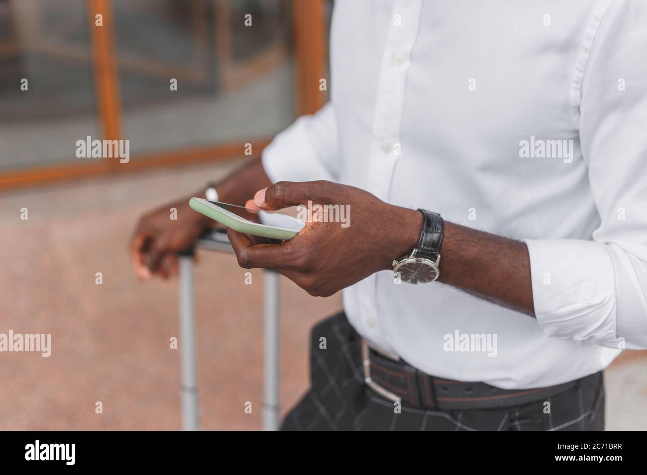 Uomo nero con borsa per bagagli in piedi sulla strada della città utilizzando smartphone, indossando abiti di stile formale Foto Stock
